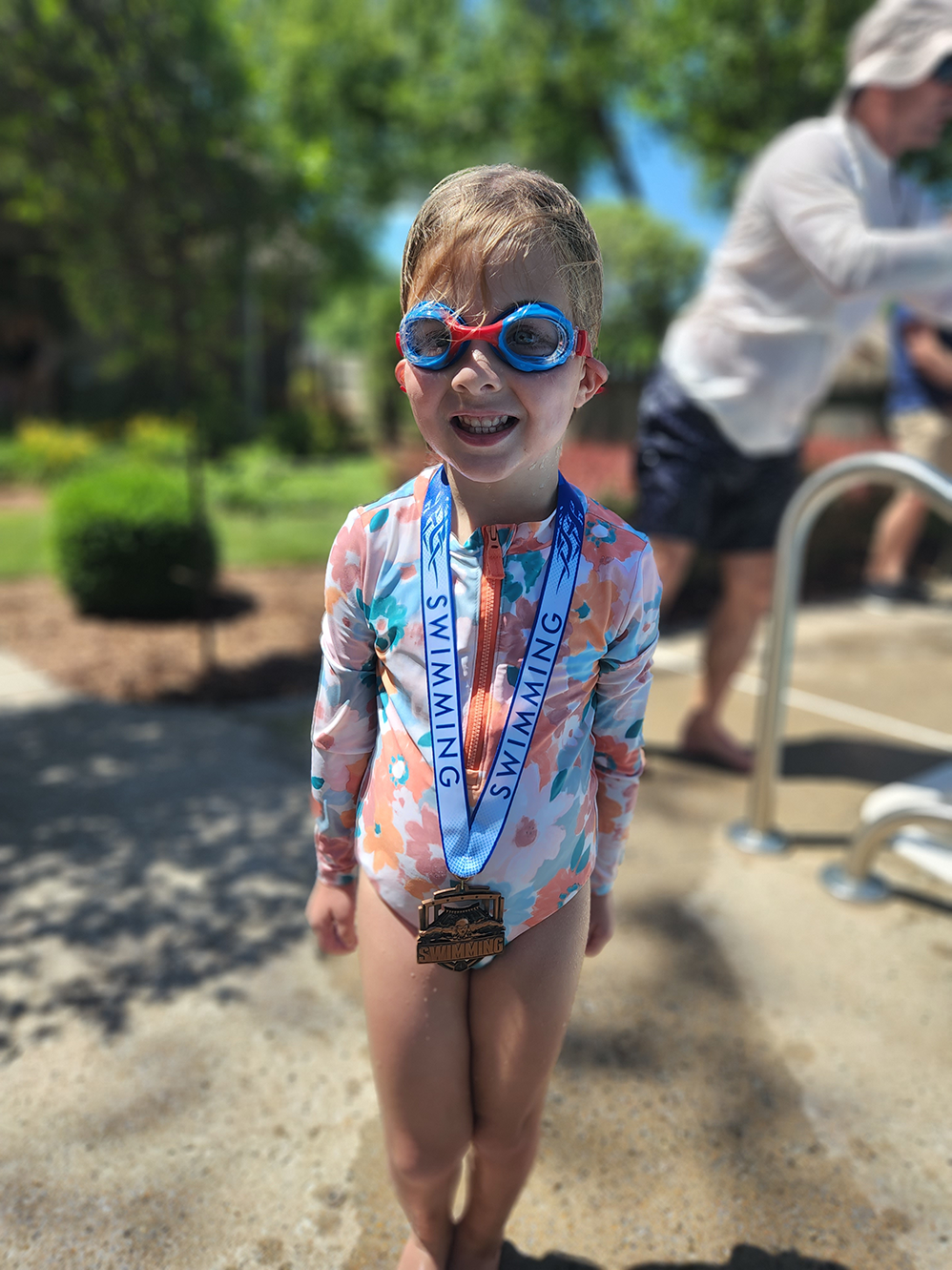 A young girl stands smiling poolside while wearing swim goggles and a completion medal at the end of her swim lessons.