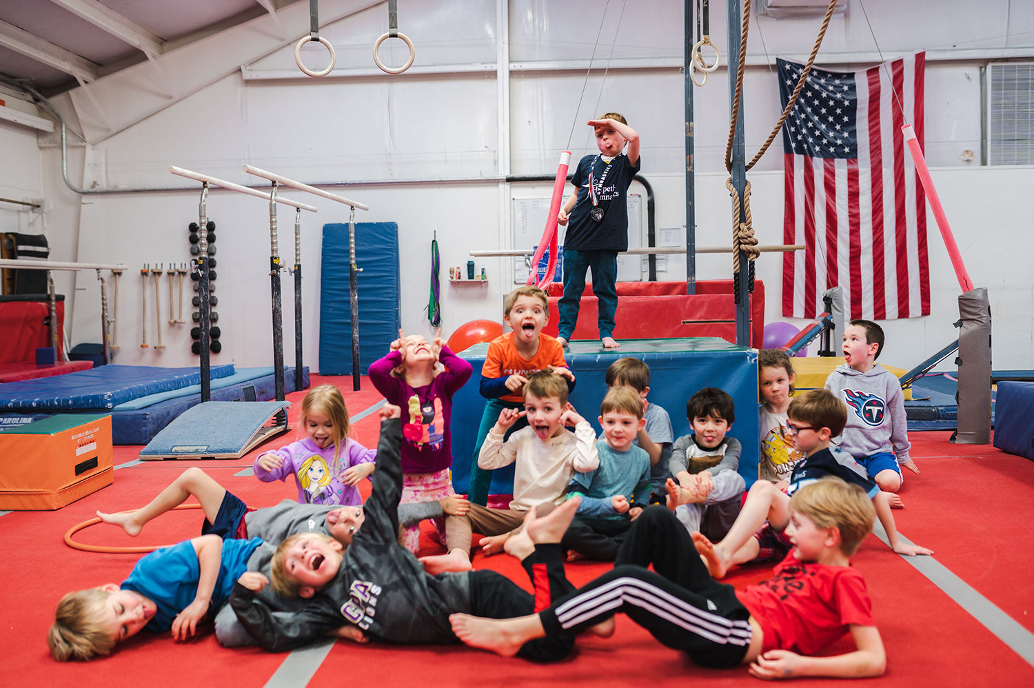 A young boy stands atop a gymnastics tumbling structure surrounded by friends who are celebrating his birthday. The children are making silly faces for the camera.
