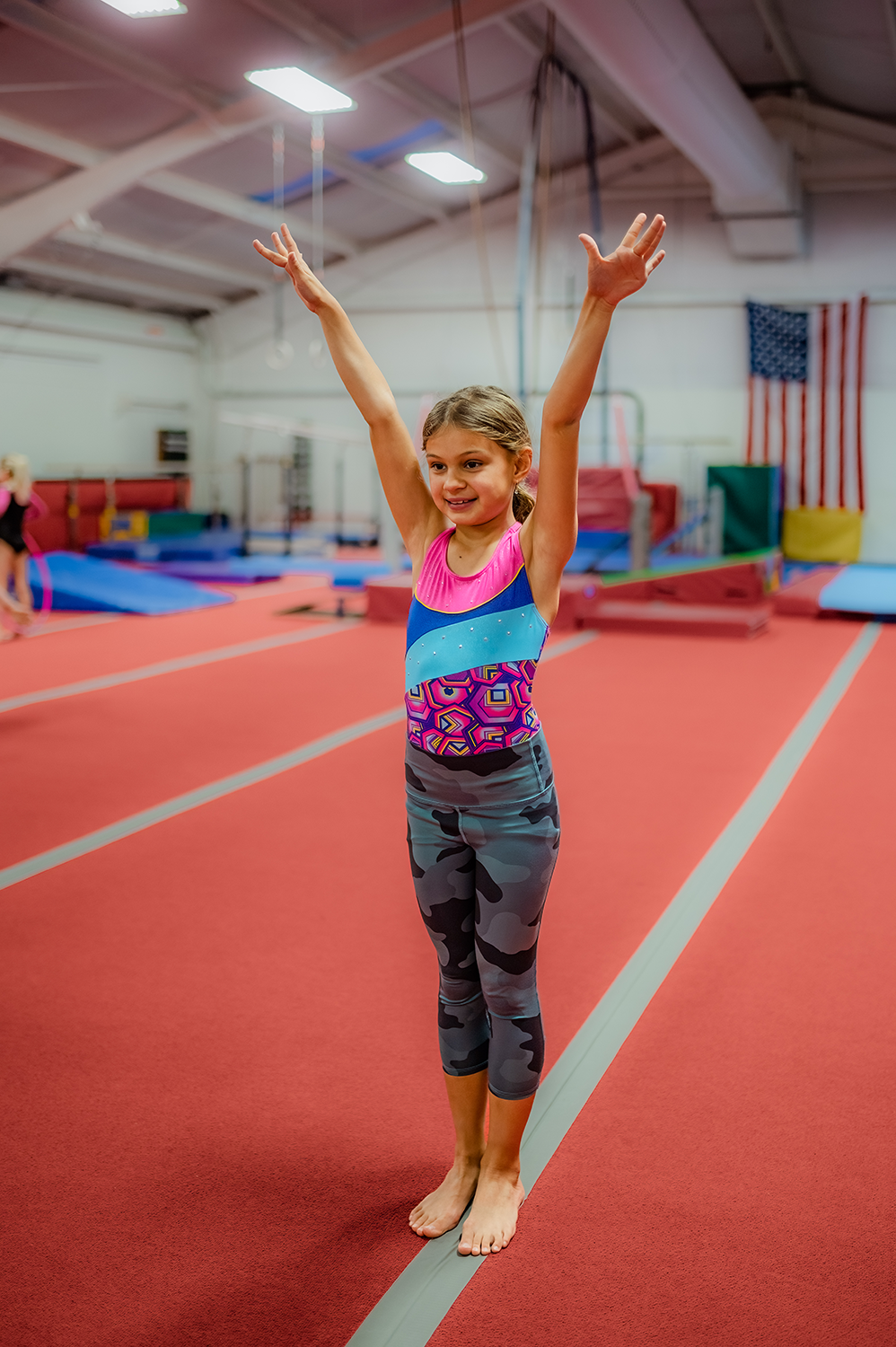 A young girl dressed in a gymnastics leotard stands with arms raised overhead practicing her final stance.