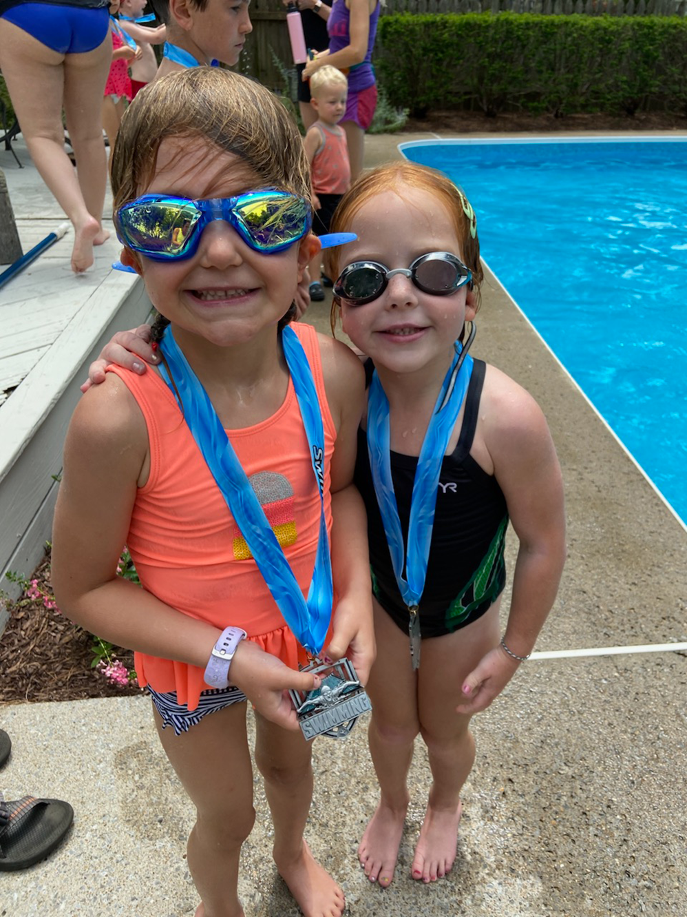 Two young girls wearing swim goggles stand poolside displaying their medals received upon completing their swim instruction.